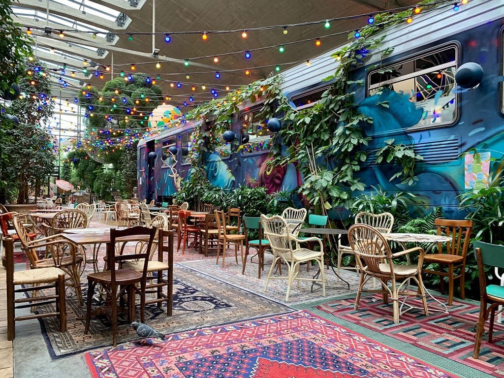 Vibrant indoor café in Paris with eclectic seating, colorful string lights, and a graffiti-covered train surrounded by greenery